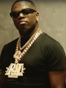 Big Boogie, wearing a black t-shirt and showcasing an extravagant gold chain necklace with a large "R4L Business" pendant adorned with diamonds, paired with diamond stud earrings. The person sports dark sunglasses, exuding a bold hip-hop vibe against a neutral background, reflecting the lavish lifestyle