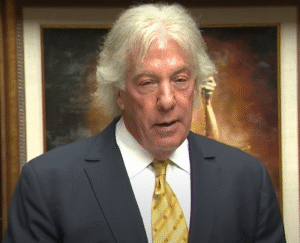 Geoffrey Fieger wearing a navy blue suit, white shirt, and yellow patterned tie, speaking in front of a framed artwork — used to represent his success and professional stature in an article about Geoffrey Fieger’s net worth.