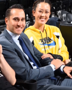 “Jonnie West smiling in a gray suit beside his wife Michelle Wie West, who is wearing a yellow Nike hoodie, during a basketball game.”