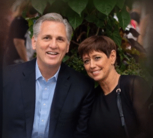 "Judy McCarthy smiling beside her husband Kevin McCarthy during an outdoor event, both dressed formally with greenery in the background."