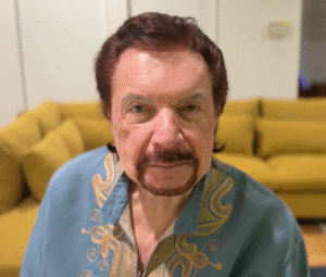 "Mike Murdock wearing a blue shirt with gold embroidered patterns, sitting in front of a yellow couch."