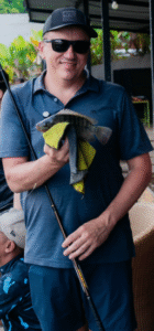 Shane Van Boening smiling while holding a fish during a fishing trip, wearing sunglasses and a blue polo shirt.