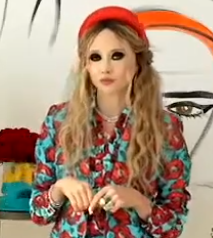 Stacey Bendet dressed in a colorful floral outfit with a red headband, standing in front of artistic wall decor.