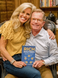 A smiling couple, Steve Houghton and Jennifer Houghton, sitting together in a cozy room with bookshelves in the background. Steve holds a copy of his book "Rich Routines: Simple Habits That Enrich Every Area of Your Life," while Jennifer rests her arm around him. She wears a yellow top with lace sleeves, and he is dressed in a light blue shirt and khaki pants.