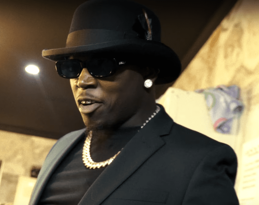 Big Boogie, wearing a black suit and stylish black fedora hat with a feather, accessorized with large diamond earrings and a chunky gold chain necklace, captured in a warm, dimly lit setting, possibly at a performance or event. Highlights the lavish lifestyle tied to Big Boogie net worth in 2025, appealing to fans of hip-hop wealth and celebrity biography