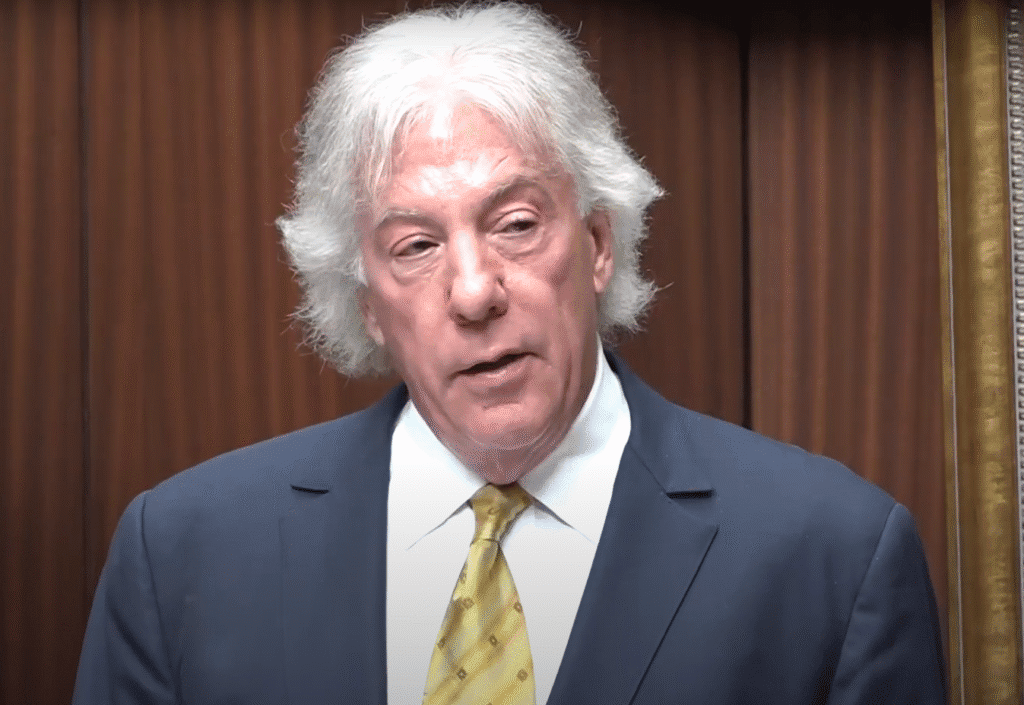 Geoffrey Fieger with white hair wearing a navy blue suit, white shirt, and yellow tie speaking in front of a wooden background.