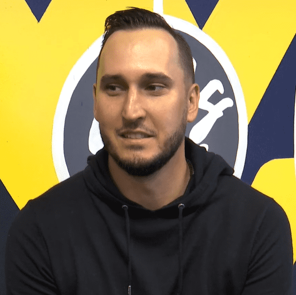 “Jonnie West wearing a black hoodie during an interview, sitting in front of a yellow and blue background.”